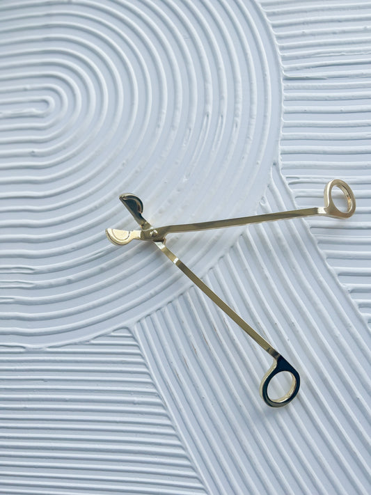 an open, gold wick trimmer on top of a decorative background.  the wick trimmer resembles slanted scissors